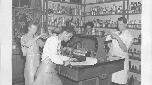 Quatre jeunes hommes sont au travail dans un laboratoire. Ils sont autour d'une table où se trouvent des instruments, deux mesurent des substances, un autre prend des notes. Autour d'eux se trouvent des étagères remplies de fioles et de bouteilles diverses. Quatre jeunes hommes sont au travail dans un laboratoire. Ils sont autour d'une table où se trouvent des instruments, deux mesurent des substances, un autre prend des notes. Autour d'eux se trouvent des étagères remplies de fioles et de bouteilles diverses.