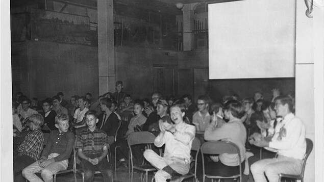 Plusieurs élèves sont assis sur des chaises en métal dans une salle et applaudissent. Il y a derrière eux un écran blanc. Plusieurs élèves sont assis sur des chaises en métal dans une salle et applaudissent. Il y a derrière eux un écran blanc.