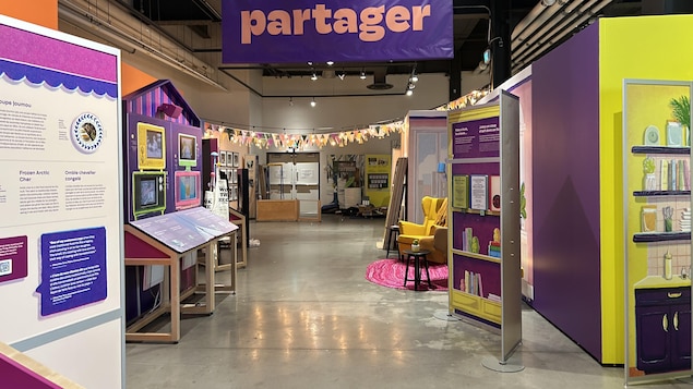 Le couloir d'une exposition de musée divisé en plusieurs sections, tous très colorées. Il est écrit ''Partager'' sur une grande affiche accrochée du plafond.  