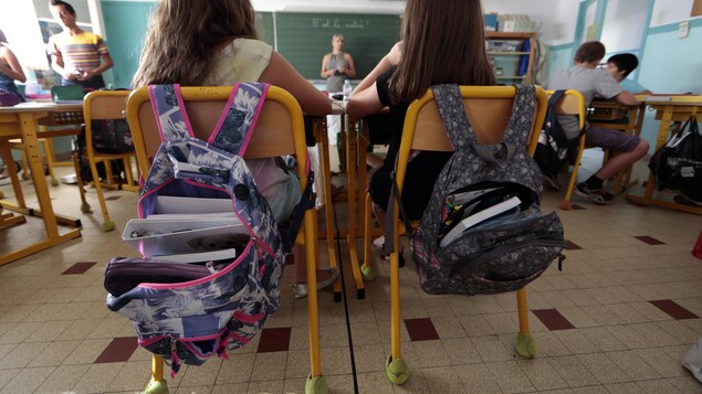 Deux petites filles photographiées de dos assises à leur place dans une classe, regardant dans la direction de leur enseignante. Leurs sacs d'école sont posés sur le dossier de leurs chaises, ils sont ouverts et on voit des livres à l'intérieur.