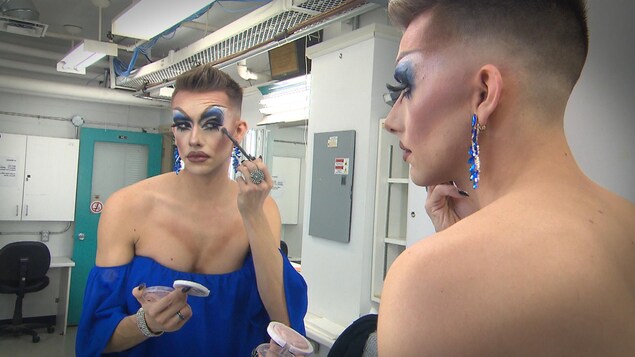 Une drag queen applique son maquillage devant un miroir d'une loge.