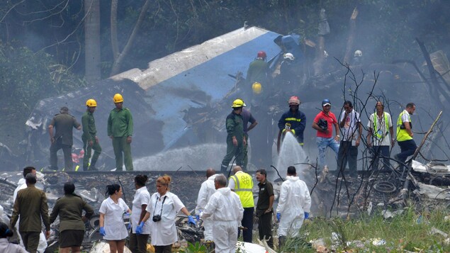 De nombreuses personnes, dont certaines qui portent un casque ou des gants, se trouvent près d'une carcasse d'avion. Une aile brûlée est tout ce que l'on voit parmi les débris. De nombreuses personnes, dont certaines qui portent un casque ou des gants, se trouvent près d'une carcasse d'avion. Une aile brûlée est tout ce que l'on voit parmi les débris.