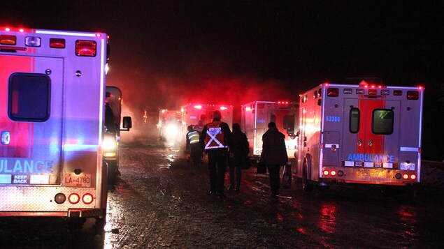 Des premiers répondants et des civils marchent sur la route à la noirceur au milieu de plusieurs ambulances à la suite d'un accident.
