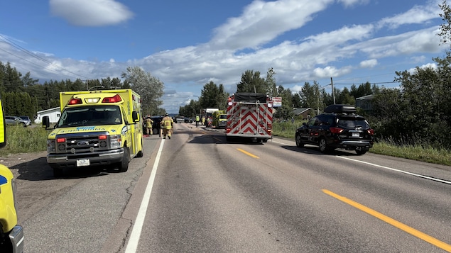 Une scène d’accident sur une route à deux voies par temps dégagé. Une ambulance jaune est garée à gauche, un camion de pompiers au centre, et des intervenants d'urgence s'activent au loin près d'un véhicule noir immobilisé à droite.