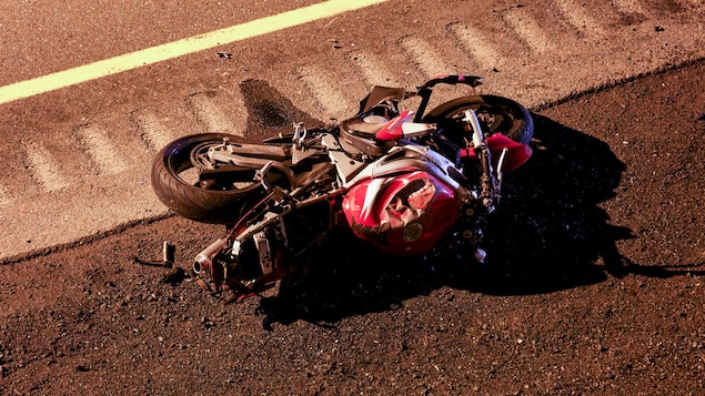 Une moto endommagée au niveau du réservoir d'essence est couchée par terre en bordure d'une route. 