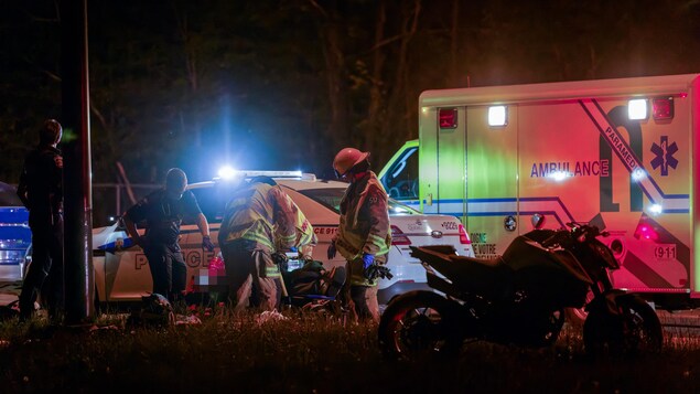 Des pompiers et ambulanicers entourent le corps d'un homme, une moto est visible dans l'obscurité