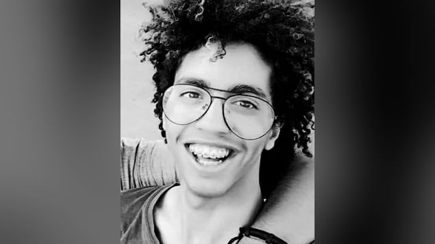 Portrait en noir et blanc d'Achraf Thimoumi, unjeune homme portant des lunettes de vision, souriant pour la caméra. Il a des broches et les cheveux bouclés.