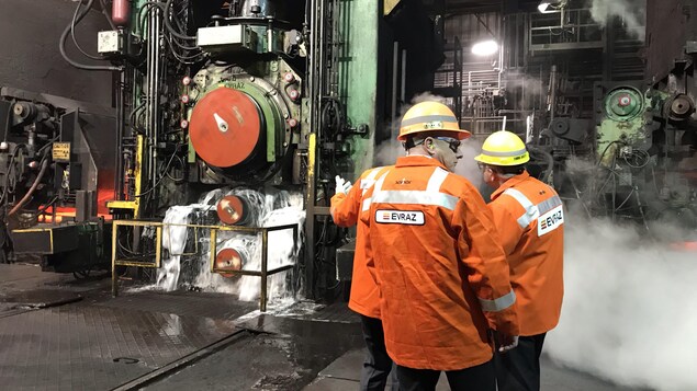 Trois hommes dans une usines portent des vestes oranges et regardent la machinerie