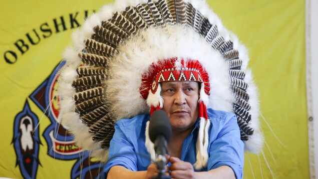 Un chef de Première Nation assis derrière un micro et portant sur sa tête un chapeau de plumes.