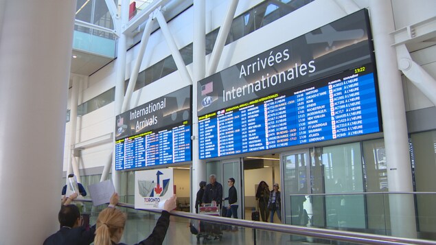 Des tableaux d'affichage des arrivées internationales à l'aéroport Pearson sous lesquels des voyageurs passent la guérite avec leurs bagages.
