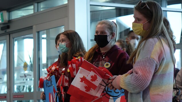 Des adolescentes qui portent le masque sanitaire et avec des drapeaux canadiens dans les mains sont dans un aéroport. 