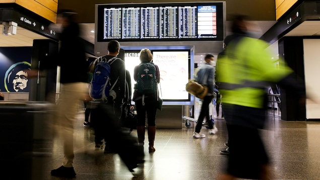 Des personnes regardent l'horaire de départ des vols sur un écran dans un aéroport.