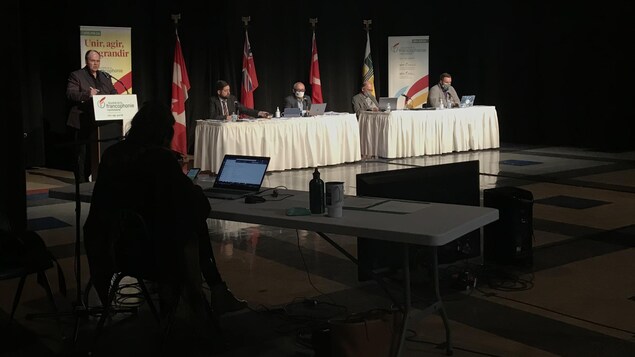 Cinq personnes espacées de deux mètres dans une grande salle. Une personne est au podium.