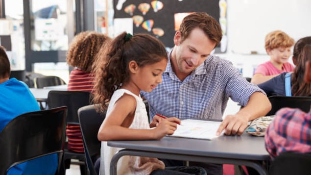 Un enseignant montre un manuel scolaire à une fillette, assise à son pupitre.