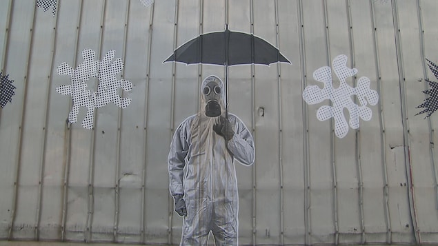 Un homme en combinaison et avec un masque et des gants, tenant un parapluie, est peint sur un mur en tôle gris.
