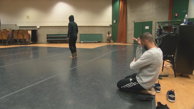 Un homme agenouillé reprend les mouvements des bras du danseur immobile, de dos, sur le plancher coussiné d'une salle de répétition. Un homme agenouillé reprend les mouvements des bras du danseur immobile, de dos, sur le plancher coussiné d'une salle de répétition.
