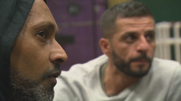 Un homme de profil sue sous un capuchon noir en avant-plan rapproché. Il regarde vers la droite, hors cadre, comme un autre homme, dont le visage est flou en arrière-plan. Un homme de profil sue sous un capuchon noir en avant-plan rapproché. Il regarde vers la droite, hors cadre, comme un autre homme, dont le visage est flou en arrière-plan.