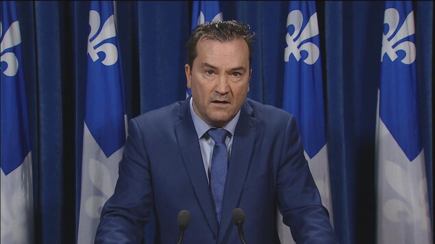 Un homme en complet, derrière deux micros, s'adresse aux médias avec l'air sérieux. Un homme en complet, derrière deux micros, s'adresse aux médias avec l'air sérieux.