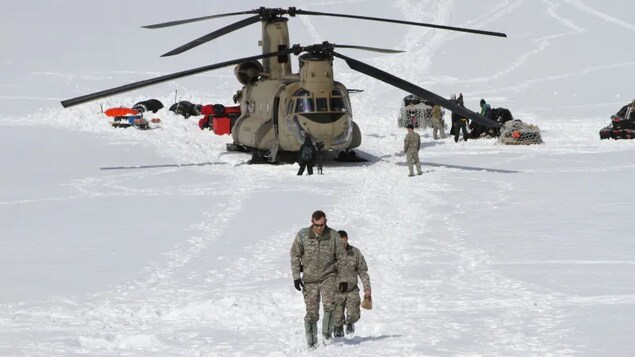 Soldiers walk across snowy terrain with a helicopter in the background. 