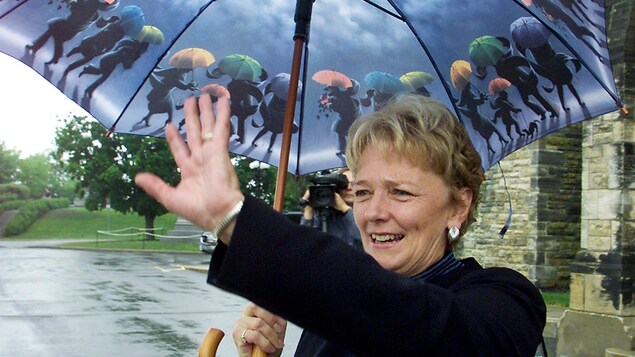 Devant le Parlement, Alexa McDonough envoie la main pour saluer en tenant de l'autre main un parapluie décoré de personnages tenant des parapluies.