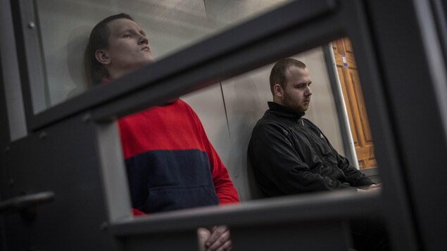 Deux hommes, le visage fermé, dans une cage vitrée. 