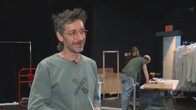 Un homme sourit devant deux personnes qui montent un décor de théâtre.