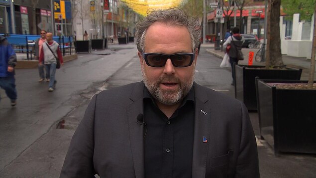 Un homme aux cheveux et à la barbe grisonnants, portant des lunettes solaires et un costume gris foncé, se tient dans une rue piétonne dans le centre-ville de Montréal et regarde droit dans la caméra.