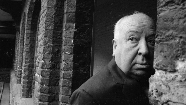 Alfred Hitchcock regarde sur le côté, alors qu'il fait face à un mur en pierre.