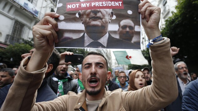 Un homme brandit une feuille sur laquelle est imprimé une photo de M. Tebboune, sur laquelle a été superposée la note "Pas mon président", en anglais.