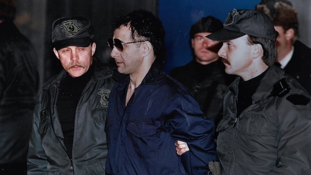Allan Legere vêtu d'une tunique bleue, fraîchement rasé et portant des verres fumés, est menotté et escorté par des policiers.