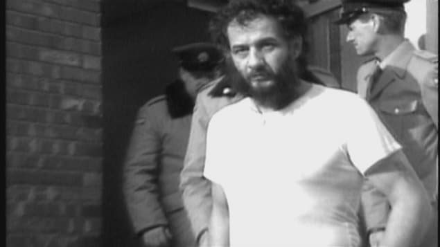 Un homme barbu sort d'un immeuble menotté et entouré de policiers.