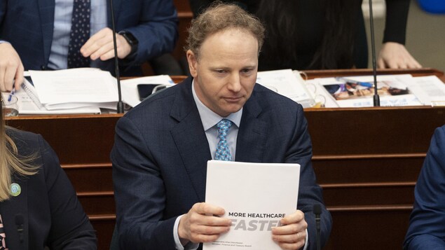 Allan MacMaster assis à l'Assemblée législative. Il est assis, il tape un document fait de plusieurs feuilles sur son bureau et regarde droit devant lui. Le nom du document est : « More Healthcare Faster ».
