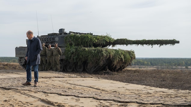 Le chancelier Olaf Scholz debout devant un char recouvert de camouflage.