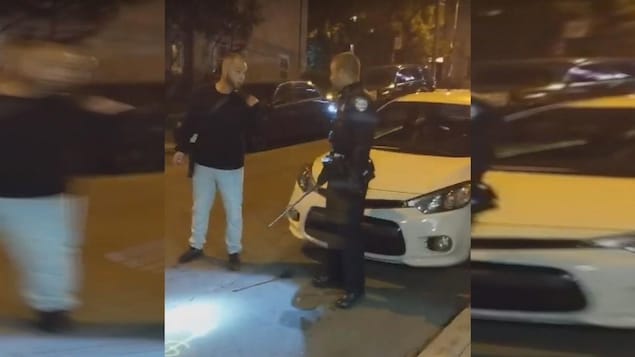 Un homme discute avec un policier tenant un bâton télescopique, devant une voiture immobilisée dans une rue.