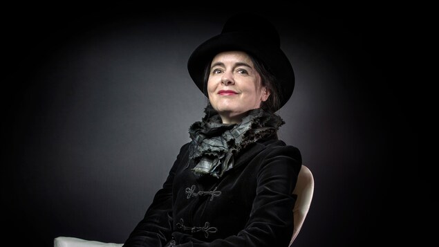 Une femme vêtue d'un veston et d'un chapeau noir prend la pose pour la caméra, assise sur un fauteuil.