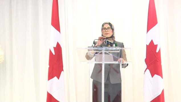 Amira Elghawaby entourée de deux drapeaux canadiens parle au micro en conférence de presse.
