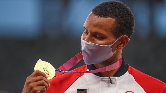 Andre De Grasse, le visage masqué, regarde sa médaille d'or.