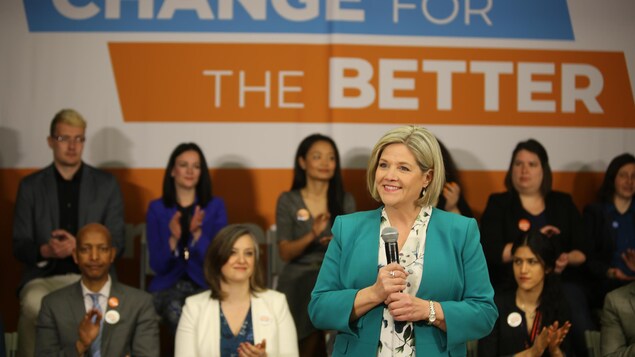 Andrea Horwath sourie, debout devant des supporteurs, avec un micro à la main. Andrea Horwath sourie, debout devant des supporteurs, avec un micro à la main.