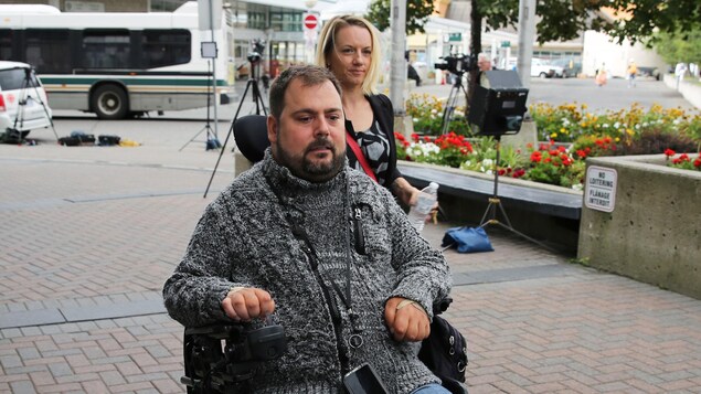 L'homme en chaise roulante suivi de près par sa conjointe et au loin plusieurs trépieds appartenant aux journalistes L'homme en chaise roulante suivi de près par sa conjointe et au loin plusieurs trépieds appartenant aux journalistes
