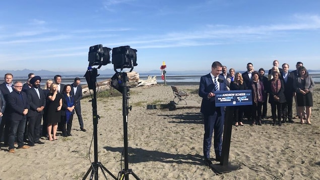 Andrew Scheer fait une annonce sur une plage en compagnie d'une vingtaine de personnes. Derriere eux, il y a de l'eau et des montagnes.