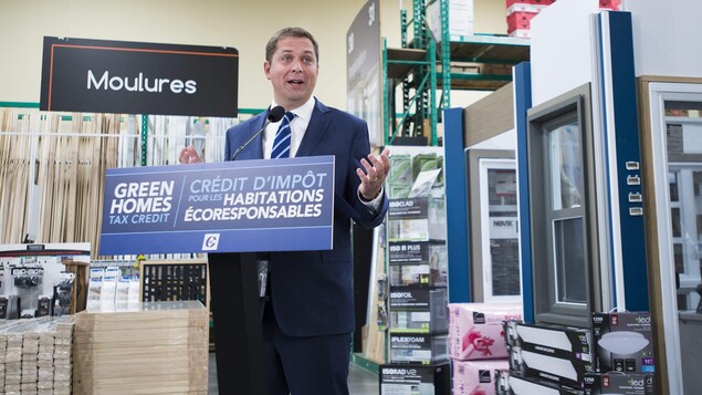 Le chef conservateur fait une annonce devant des planches de bois, dans une quincaillerie.