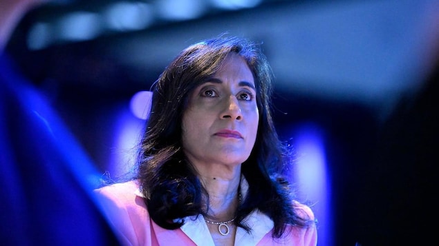 Anita Anand regarde au loin, baignée de lumière bleue dans une salle de conférence.