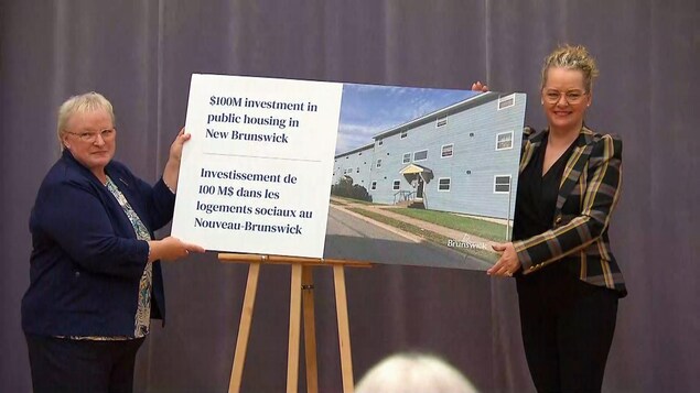 الوزيرة المسؤولة عن التنمية الاقتصادية والمؤسسات الصغيرة في حكومة نيو/نوفو برونزويك، أرلين دان (إلى اليمين) وزميلتها وزيرة التنمية الاجتماعية، دوروثي شيبارد، تحملان ملصقاً يعلن عن قرار الحكومة بناء 380 مسكناً اجتماعياً جديداً.