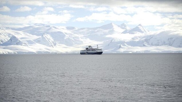 A ship shown in the water with snow covered mountains behind it. 