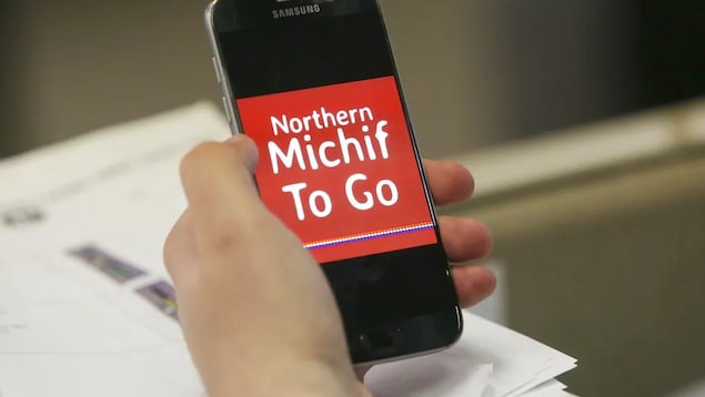 Un téléphone tenu dans une main, qui affiche l'inscription Northern Michif To Go en blanc sur fond rouge.