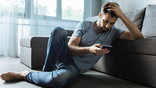 Un homme triste consulte son téléphone. Il est assis sur le tapis de son salon consulte son cellulaire.