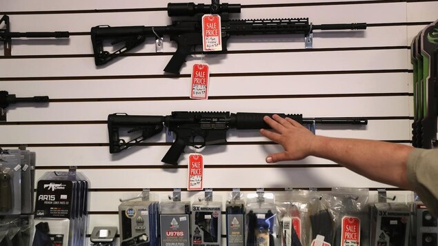 A person gestures towards assault rifles for sales mounted on a wall in a gun shop.