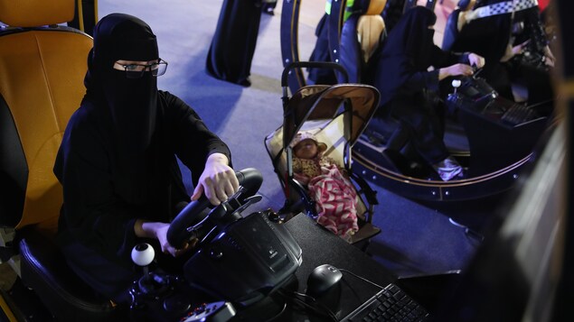 Une jeune femme portant un niqab utilise un simulateur de conduite automobile à côté d'un bébé dans une poussette. Une jeune femme portant un niqab utilise un simulateur de conduite automobile à côté d'un bébé dans une poussette.