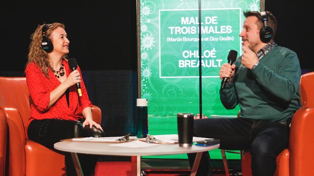 Une femme et un homme, portant des écouteurs et tenant un micro, sont assis dans des fauteuils autour d'une table de salon.