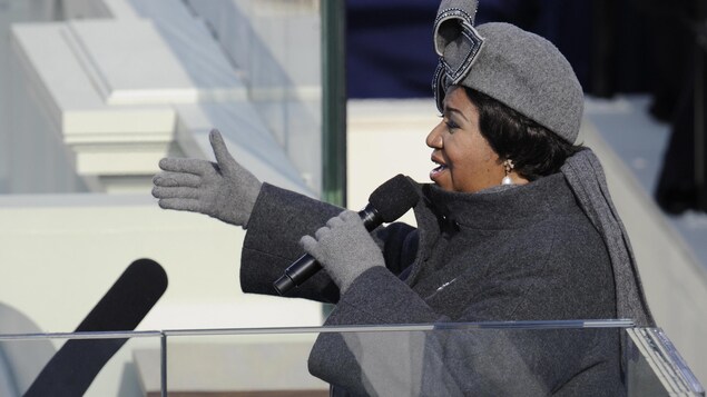La chanteuse, vêtue de gris, est de profil avec un microphone dans la main. La chanteuse, vêtue de gris, est de profil avec un microphone dans la main.